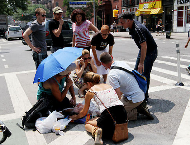 People surround a woman lying prone on a city street, shading her with a blue umbrella