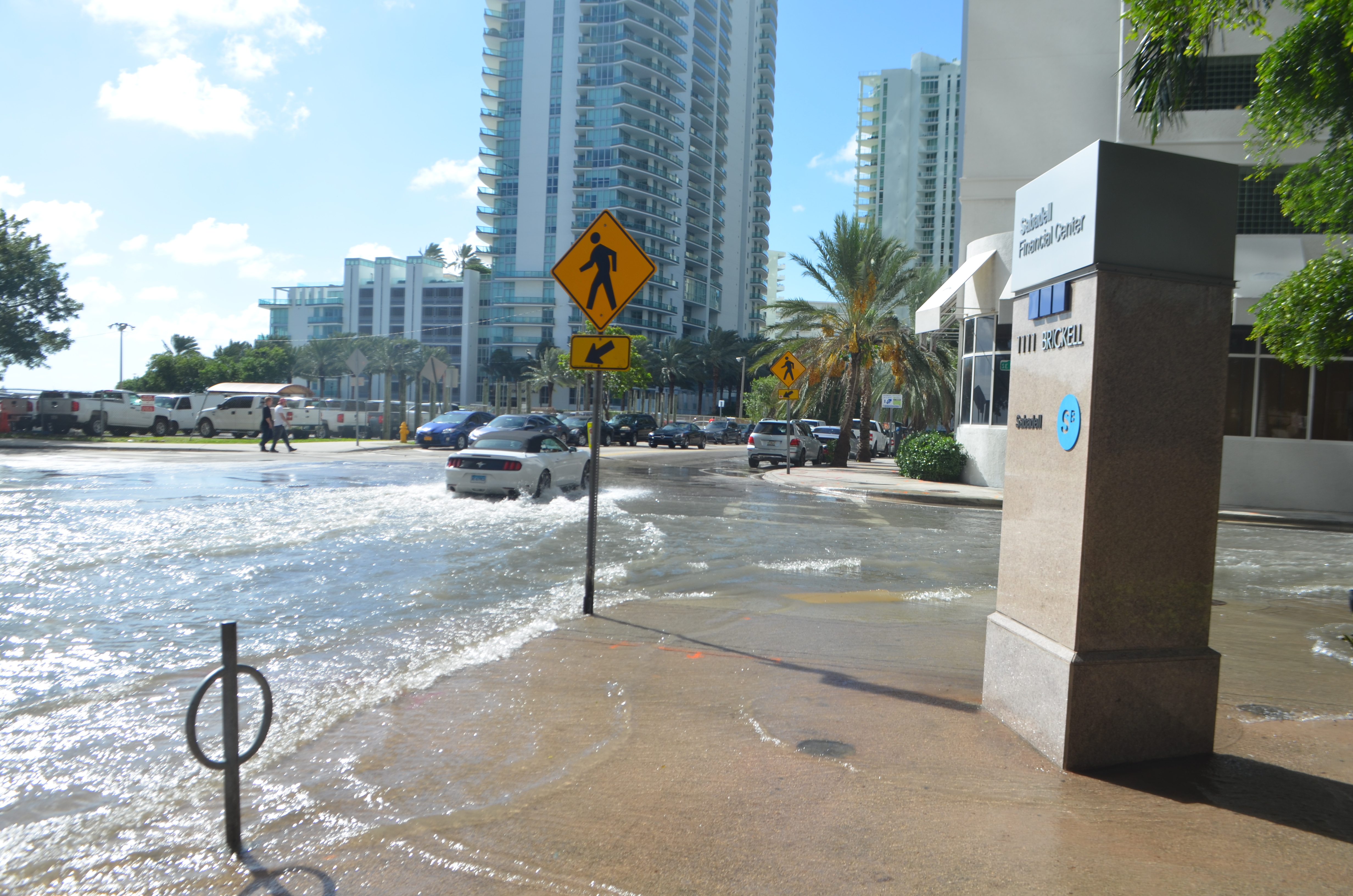 A road on left, covered in sea water, is being driven through by a car. The sun is shining indicating that the water did not fall as rain but is instead due to the tides.