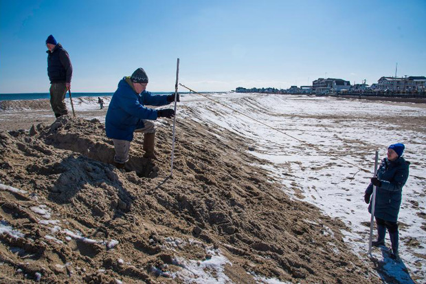 A volunteer stands on a large frozen berm on a broad beach, holding a pole attached to a measuring rope. Another volunteer holds a second pole at the inland foot of the berm.