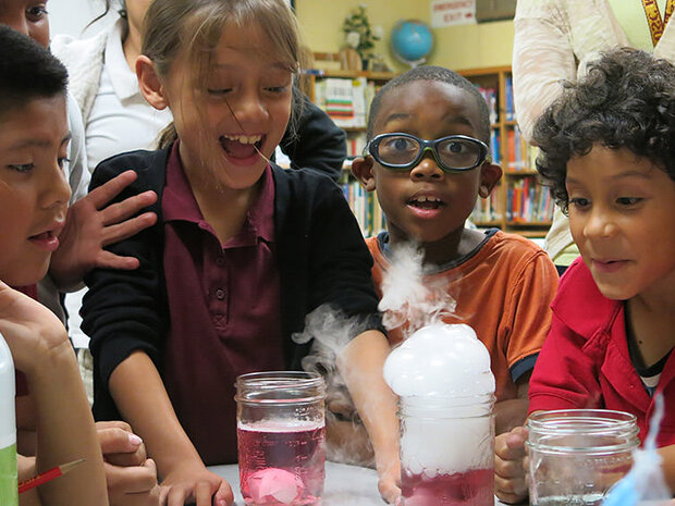 A group of students gather around a bubbling, smoking science experiment that is contained in a mason jar. The students look especially excited about the reaction happening in front of them.