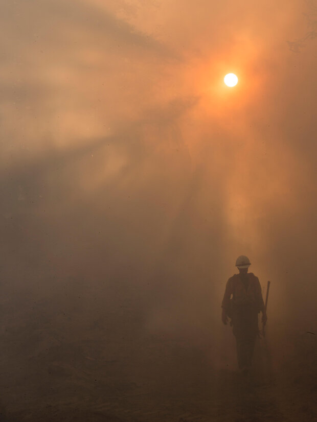 A firefighter walks into a smoke-filled area with hazy sunlight ahead of them.