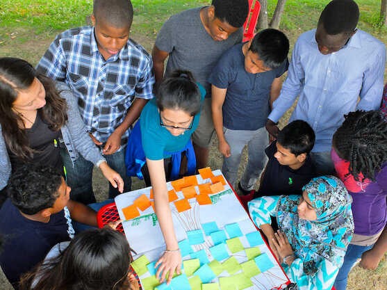 Outside in a park, an educator and students gather around an activity board with different colored sticky notes.